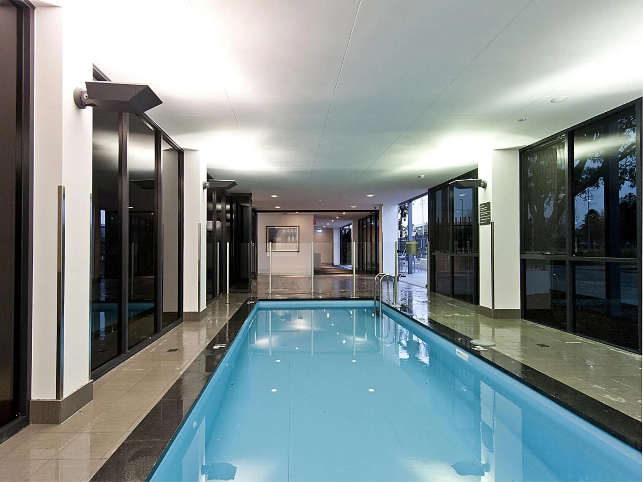 Une piscine intérieure rectangulaire avec un carrelage sombre et des baies vitrées.