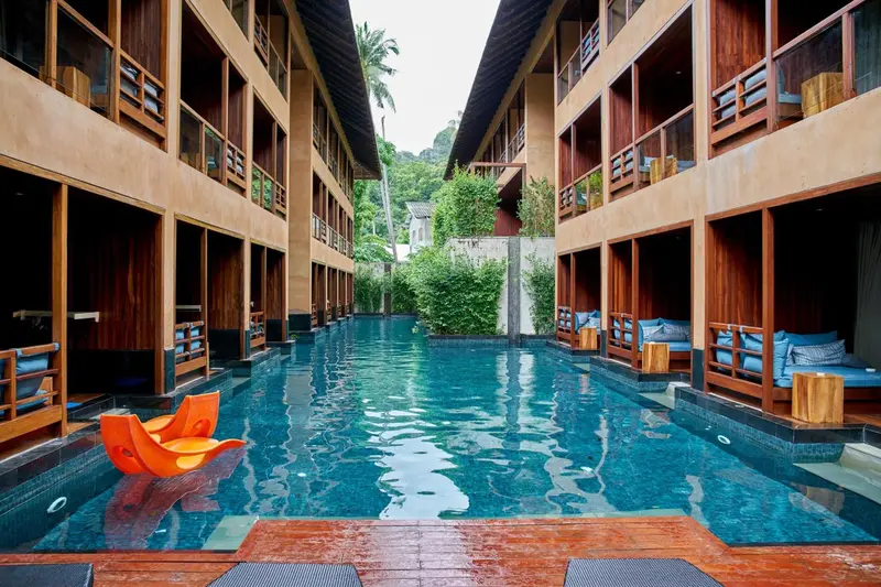 Vue d'une piscine turquoise bordée de transats et de végétation luxuriante entre deux bâtiments.