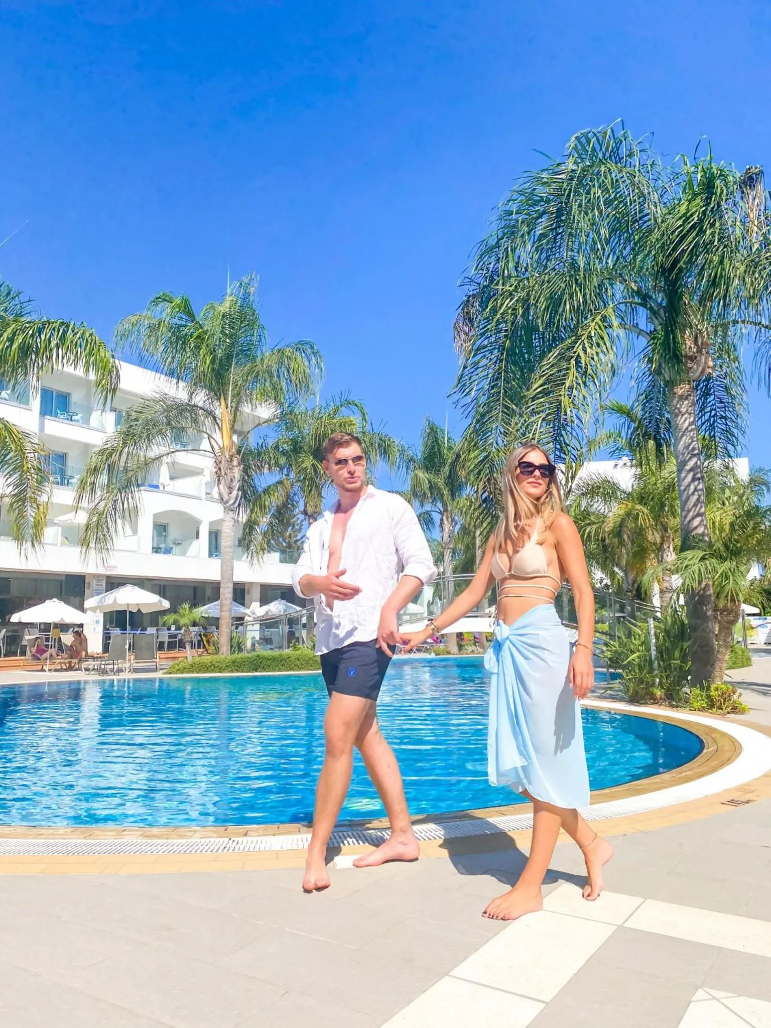 Un couple se promène le long d'une piscine turquoise bordée de palmiers.