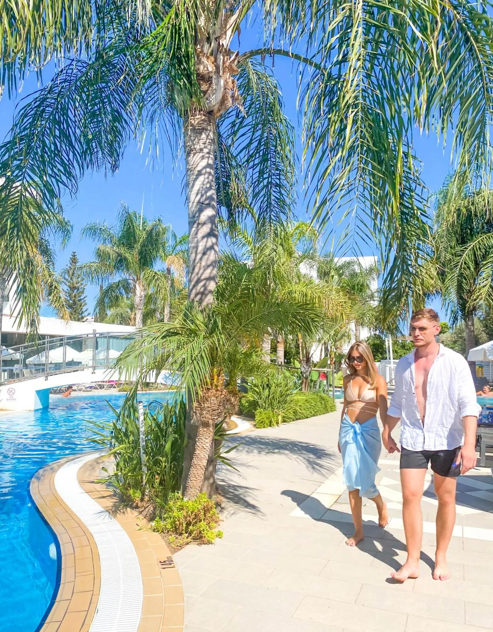 Un couple se promène le long d'une piscine bordée de palmiers.