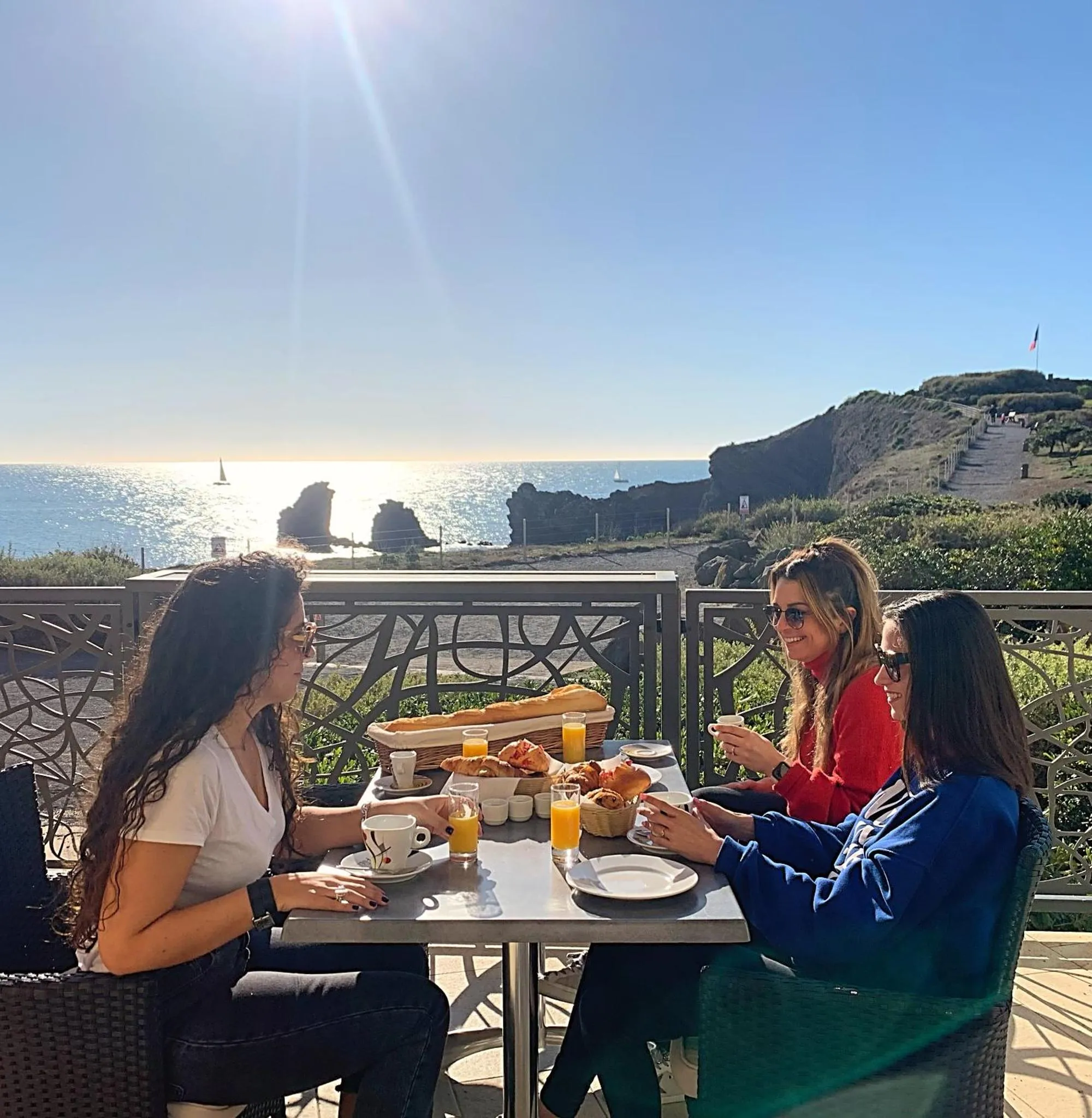 Des personnes prennent leur petit-déjeuner à une table sur une terrasse ensoleillée avec vue sur la mer.
