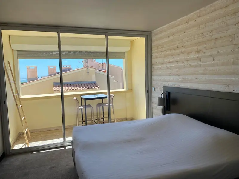Une chambre lumineuse s'ouvre sur un balcon avec une table et des tabourets.