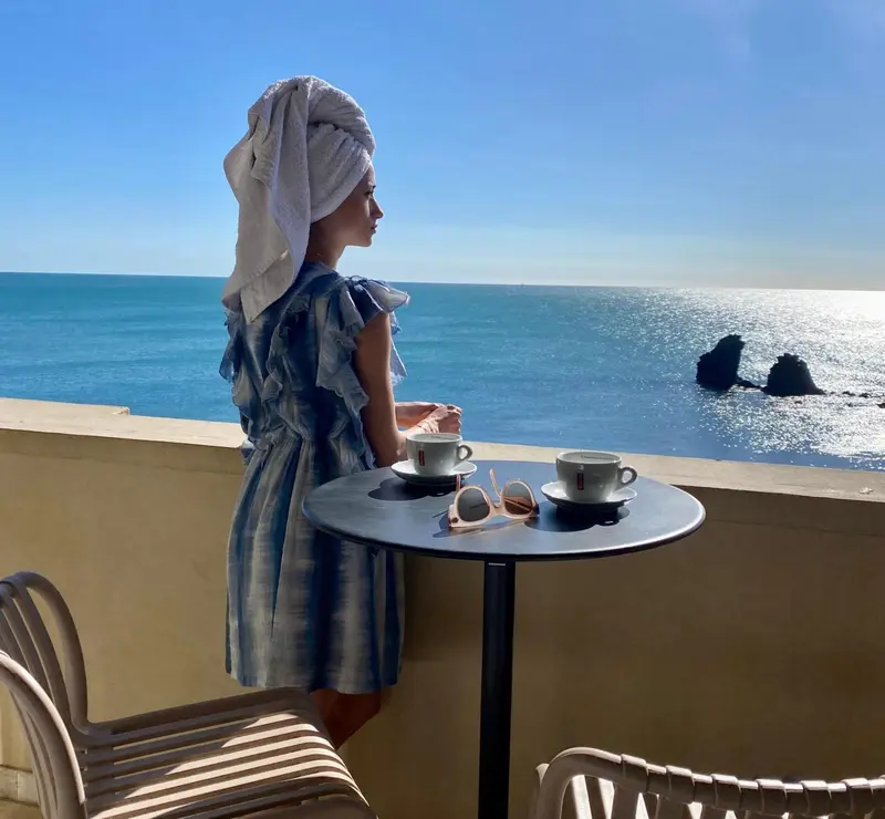 Une femme se tient sur une terrasse ensoleillée, face à la mer.