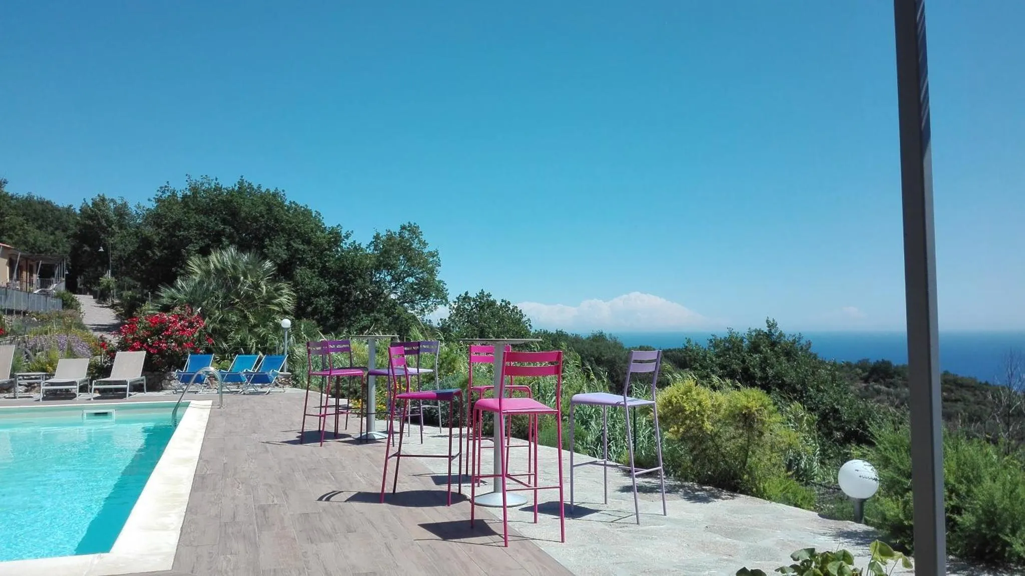 Terrasse avec chaises colorées et piscine au premier plan, végétation et vue sur la mer en arrière-plan.