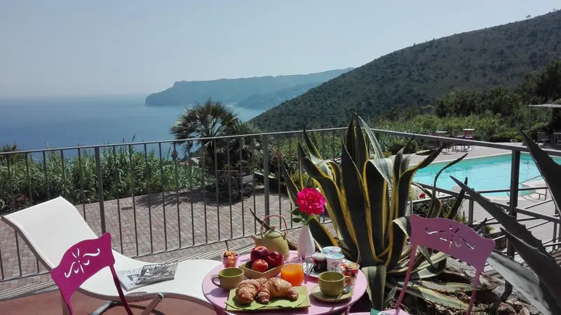 Une table de petit-déjeuner est mise en scène sur une terrasse avec vue sur la mer et les collines.