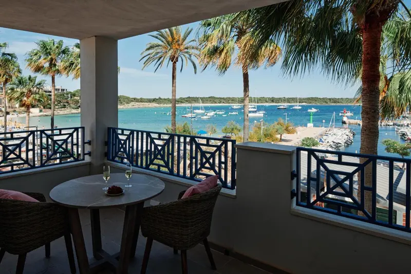 Une table ronde avec deux chaises sur une terrasse ensoleillée donne sur une baie avec des bateaux.