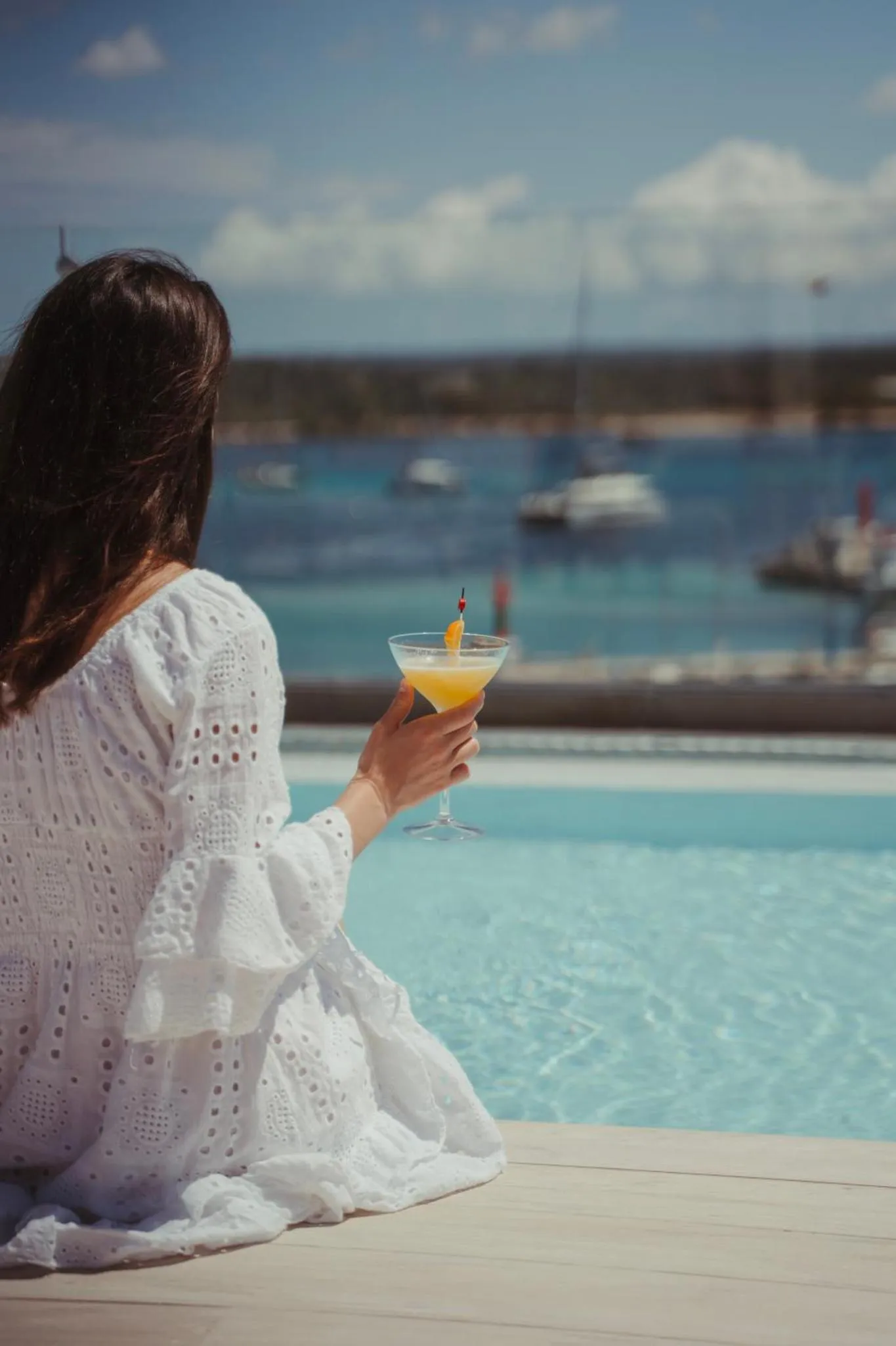 Une personne assise au bord d'une piscine, dos à la caméra, tenant un cocktail.