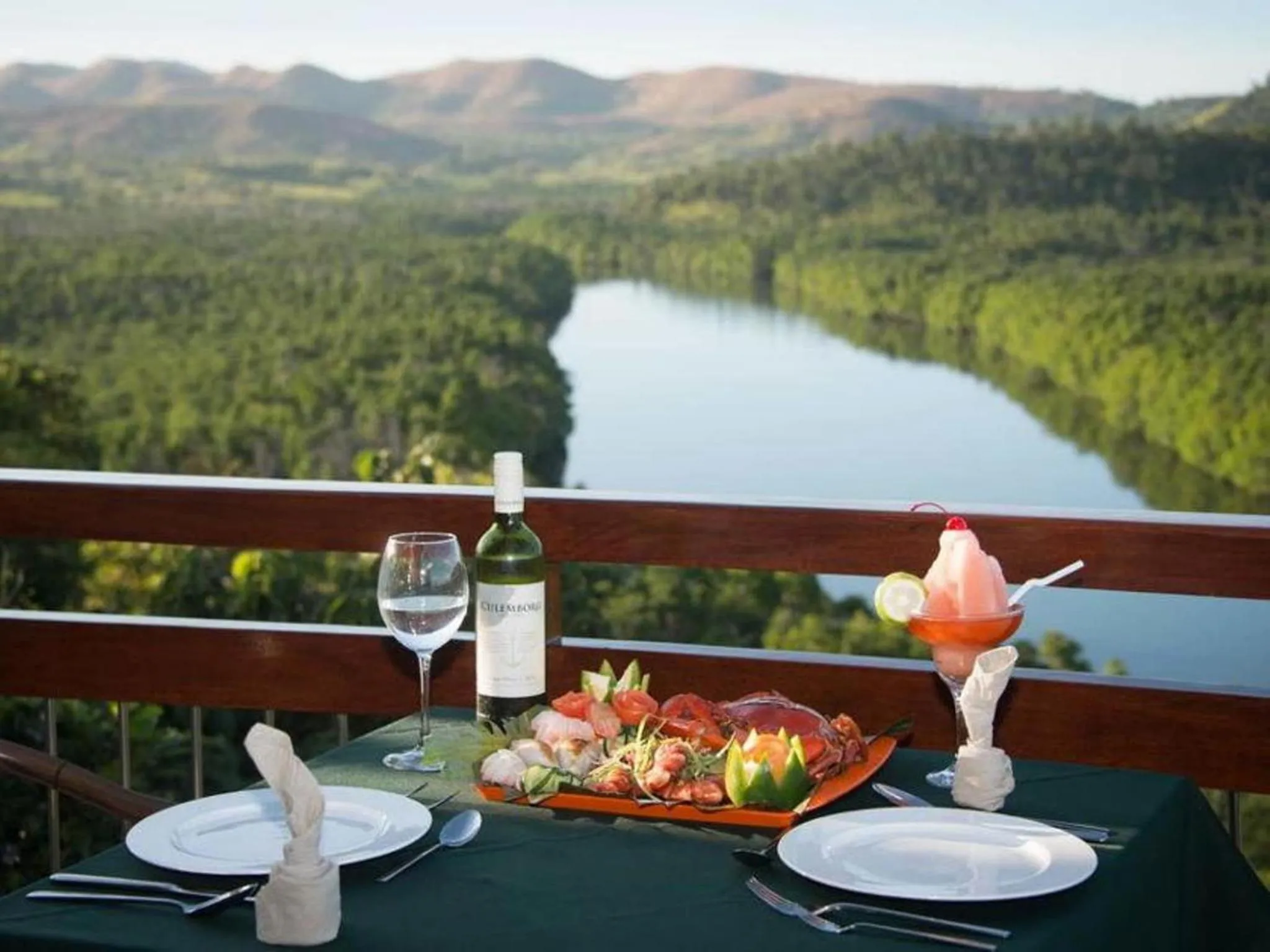 Table dressée avec des plats et des boissons sur une terrasse avec vue sur un paysage montagneux et une rivière.