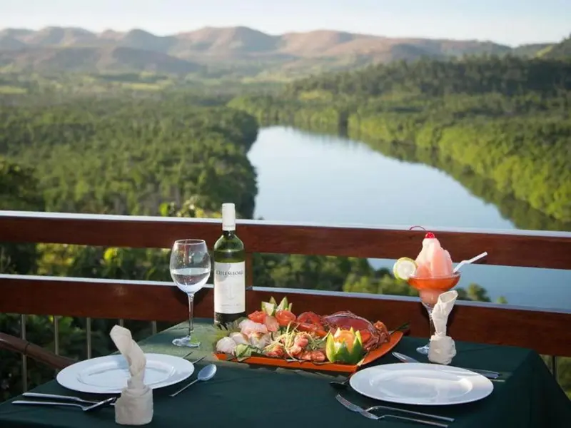 Table dressée avec des plats et des boissons sur une terrasse avec vue sur un paysage montagneux et une rivière.