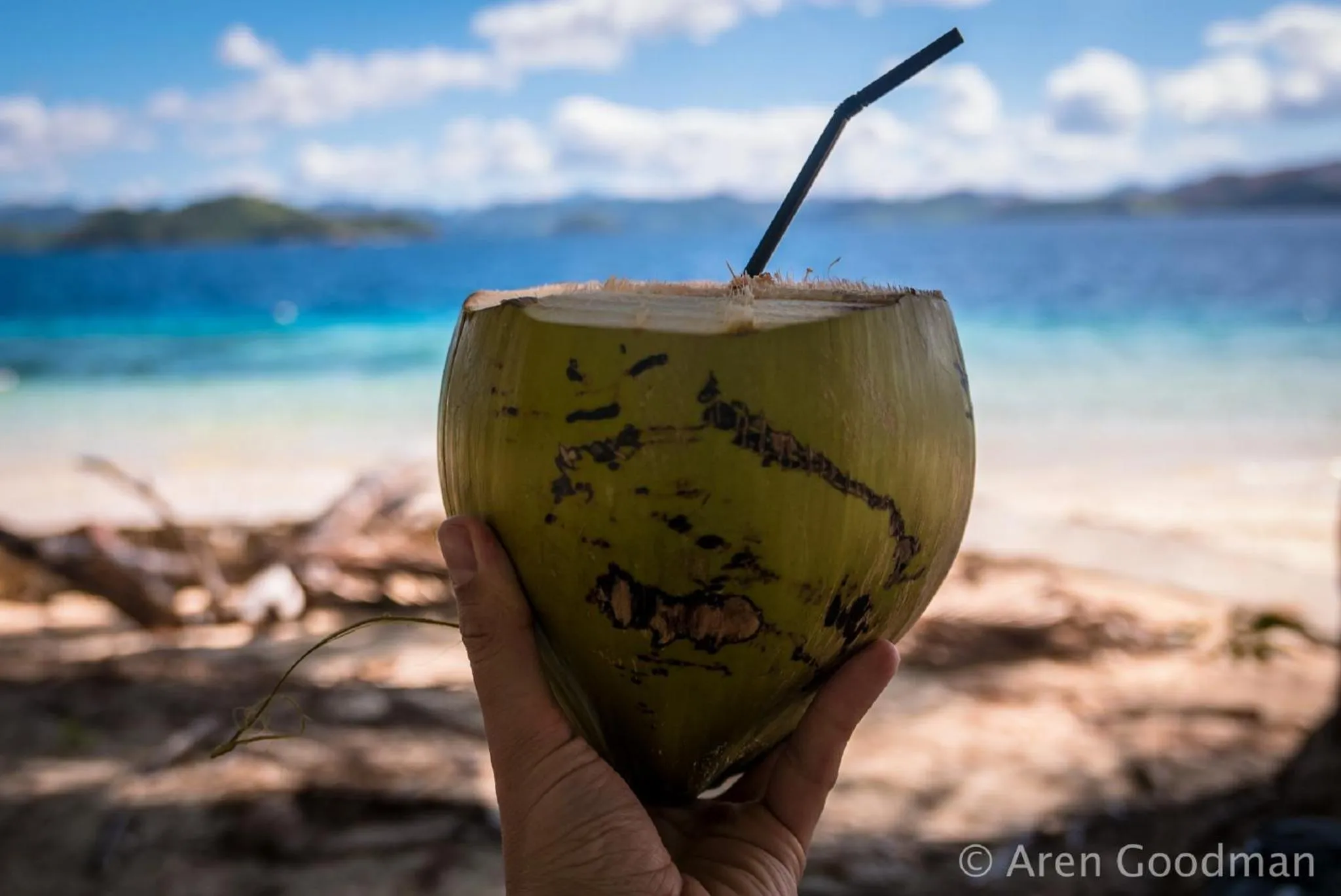 Une noix de coco fraîche coupée, tenue à la main, avec une plage et la mer en arrière-plan.