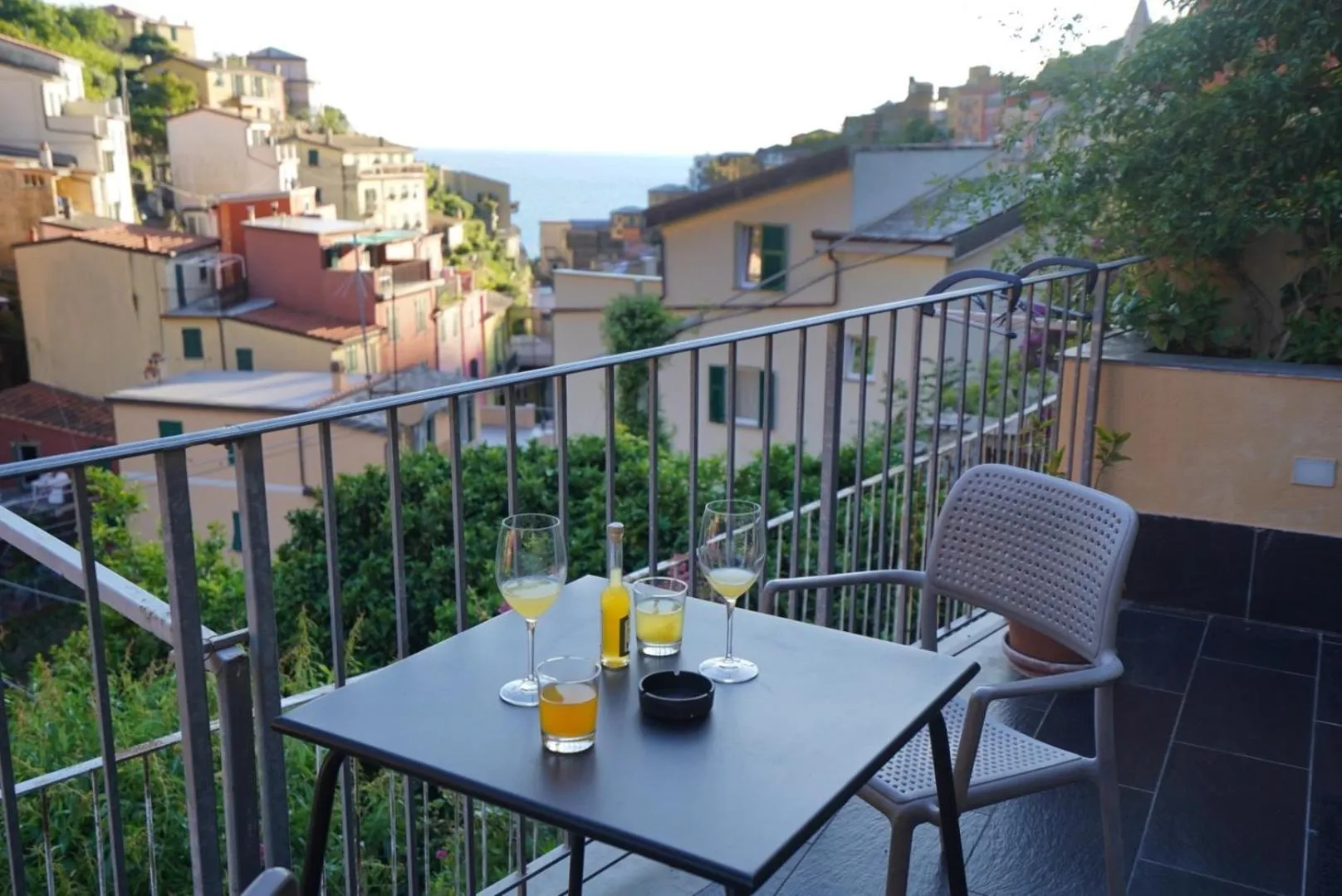 Une table avec des verres et des boissons est posée sur une terrasse ensoleillée avec vue sur la mer et les bâtiments colorés.