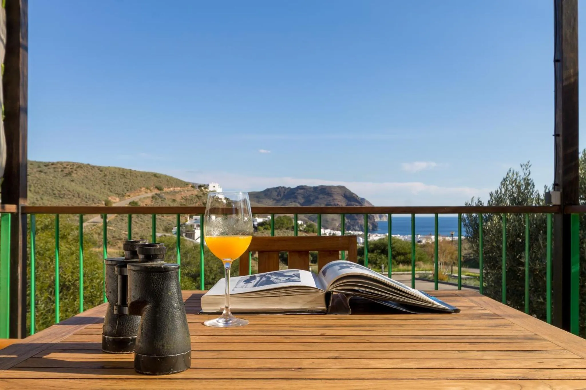 Une table en bois avec un livre ouvert et un verre de jus d'orange sur une terrasse ensoleillée.