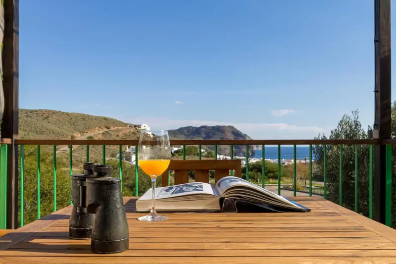 Une table en bois avec un livre ouvert et un verre de jus d'orange sur une terrasse ensoleillée.