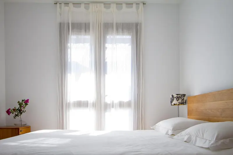 Une chambre lumineuse avec un lit recouvert de draps blancs et une table de chevet en bois.