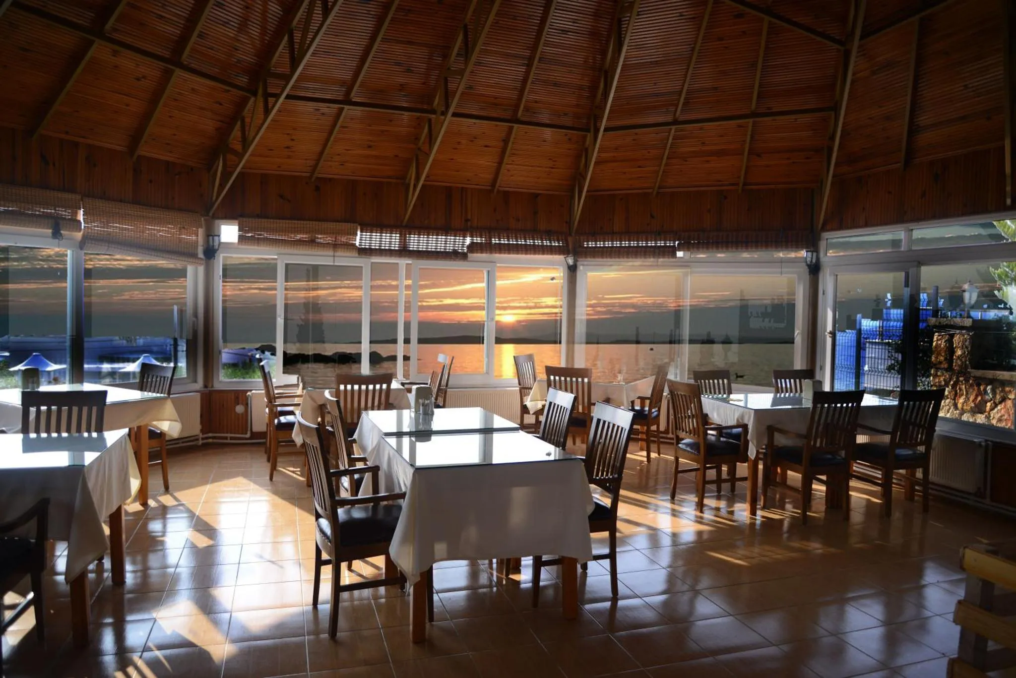 Une salle lumineuse avec des tables dressées face à un coucher de soleil sur la mer.