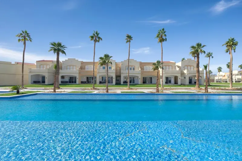 Piscine turquoise devant un bâtiment blanc bordé de palmiers sous un ciel bleu.
