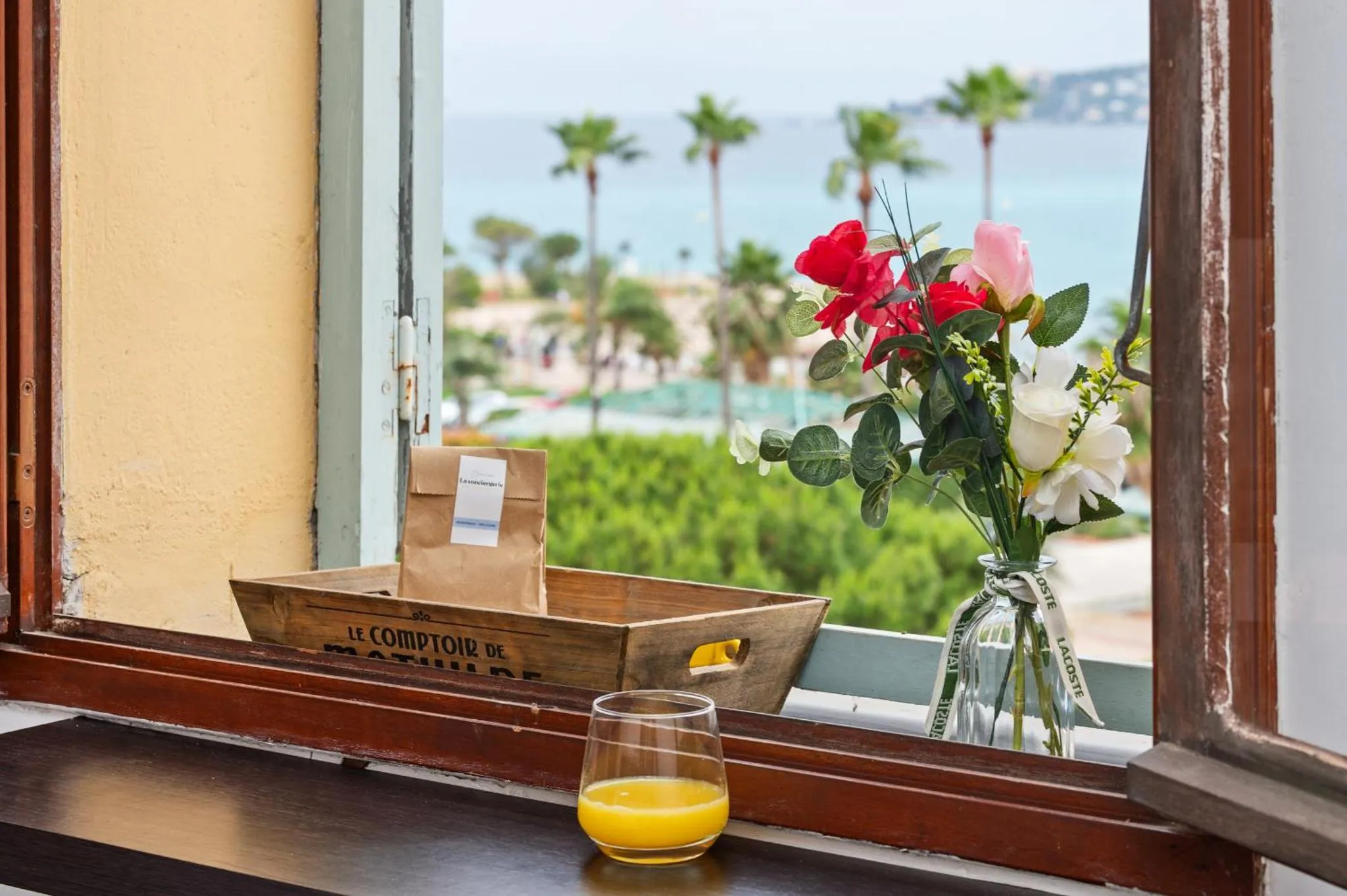 Vue sur la mer depuis une fenêtre avec un bouquet de fleurs et un verre de jus d'orange.