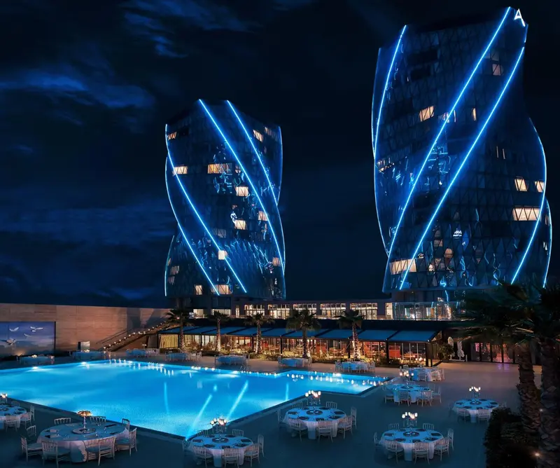 Deux bâtiments modernes illuminés en bleu se dressent au-dessus d'une piscine et d'une terrasse animée.