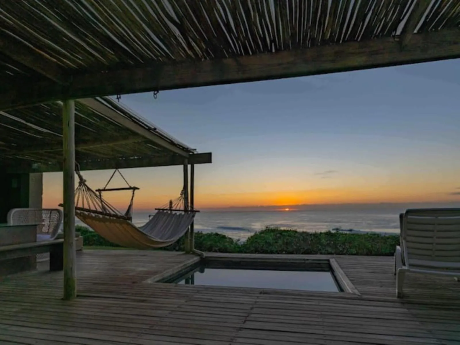 Terrasse en bois avec hamac et petite piscine face à la mer au coucher du soleil.