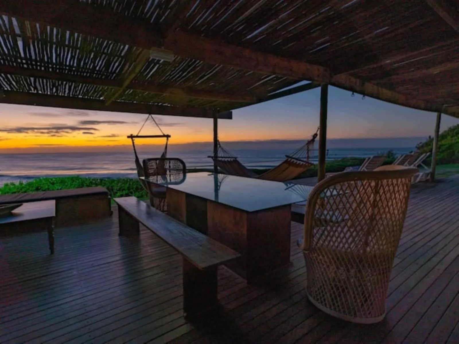 Terrasse en bois avec hamacs et mobilier de jardin face à la mer au coucher du soleil.