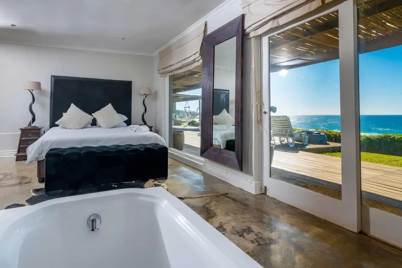 Une chambre lumineuse avec un grand lit et une baignoire, donnant sur une terrasse en bois et la mer.