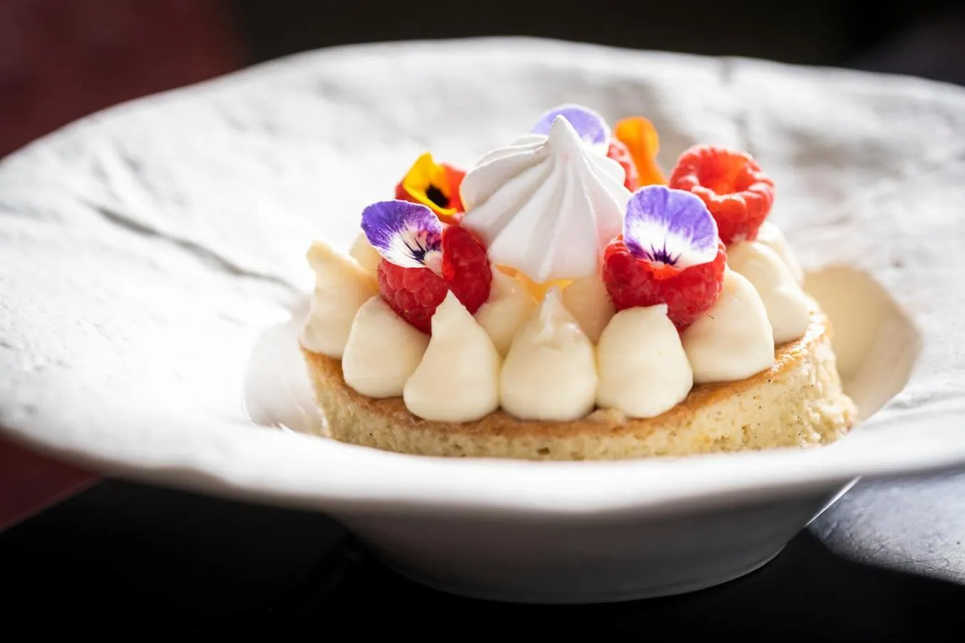 Une tartelette décorée de crème, de framboises et de fleurs comestibles est présentée sur une assiette blanche.