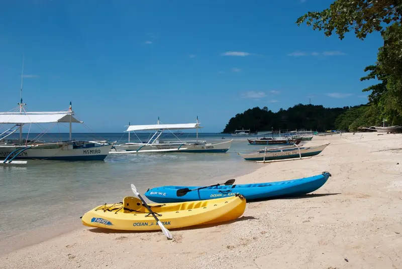 Plage de sable blanc avec des kayaks et des bateaux amarrés dans une eau turquoise.