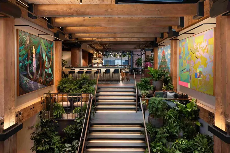 Un escalier en bois mène à un bar lumineux décoré de tableaux et de plantes.