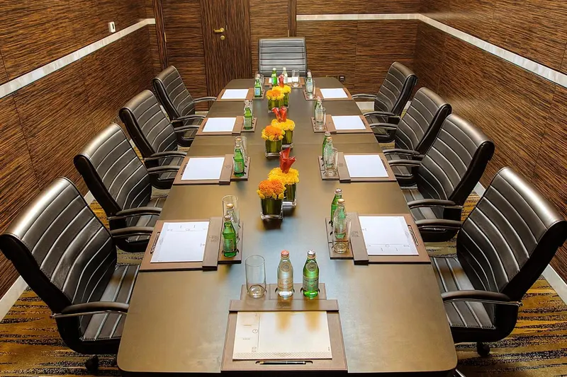 Une longue table de réunion est entourée de chaises en cuir noir, avec des bouteilles d'eau et des fleurs comme centres de table.