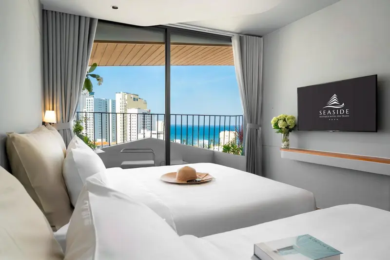 Chambre lumineuse avec un lit blanc et une vue sur la mer depuis un balcon.