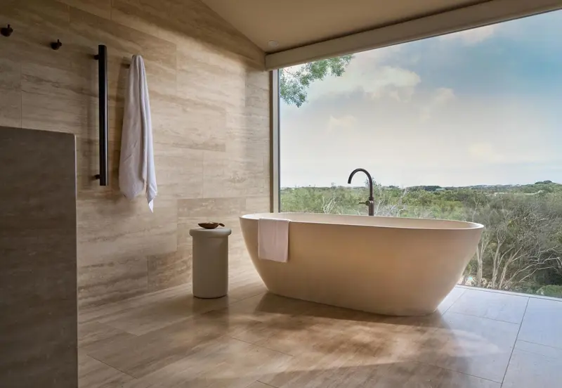Une baignoire blanche se trouve dans une salle de bain carrelée, avec une grande baie vitrée donnant sur un paysage verdoyant.
