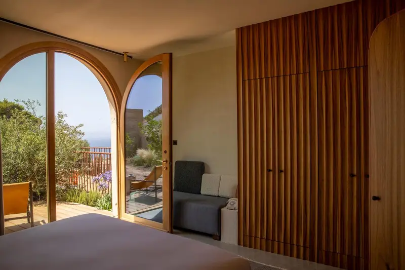 Une chambre lumineuse s'ouvre sur une terrasse avec vue sur la végétation et la mer.