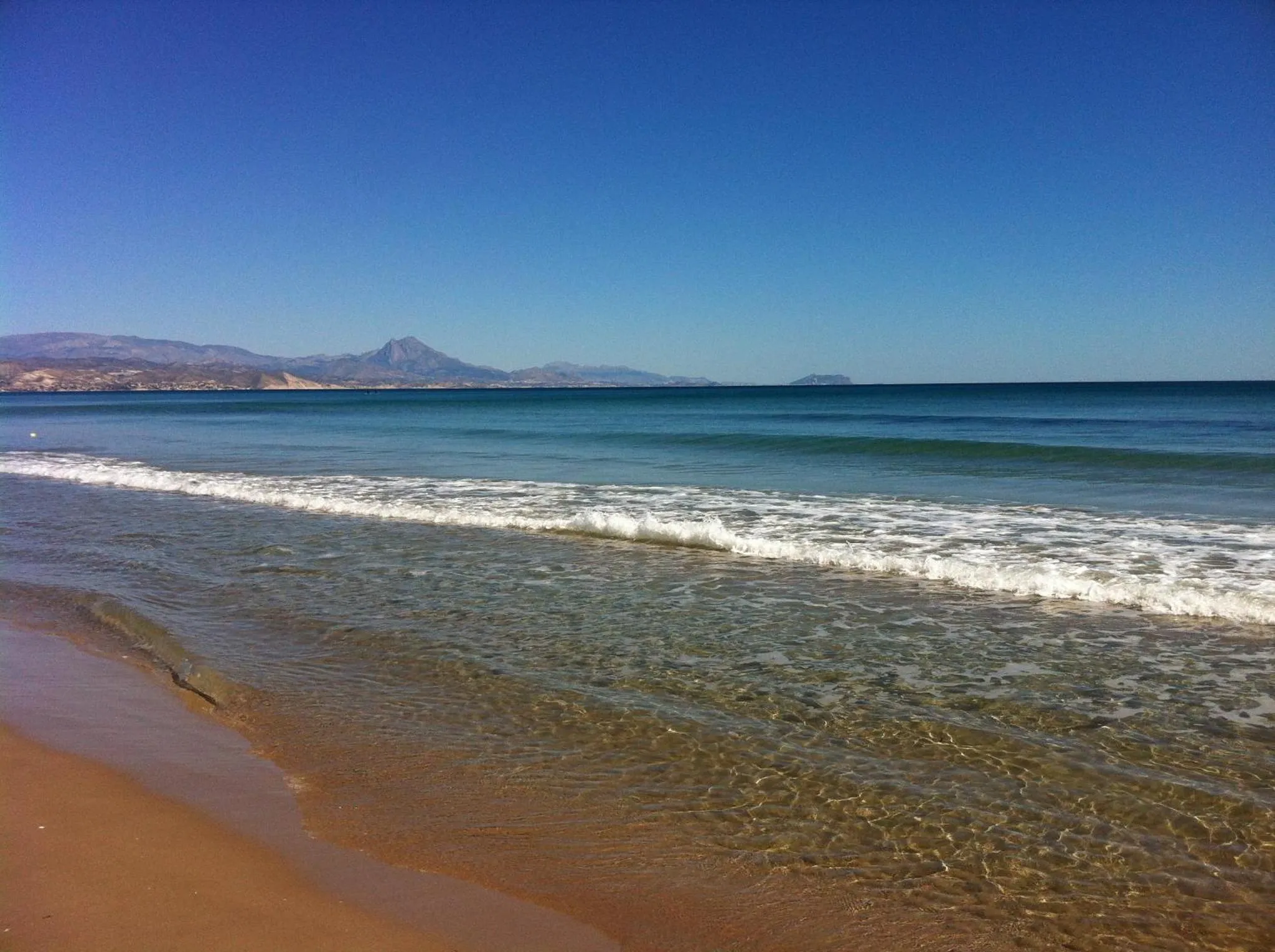 Vague déferlant sur une plage de sable fin avec une vue sur la mer et les montagnes.