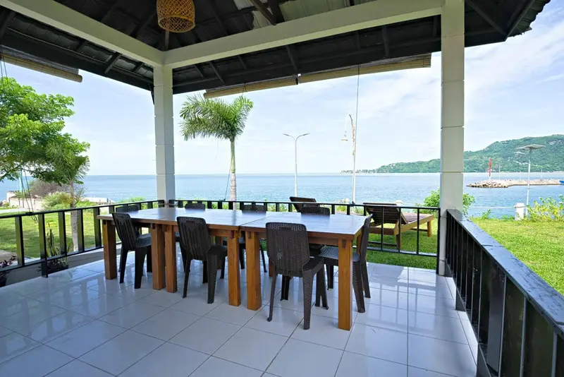 Une longue table en bois est entourée de chaises sur une terrasse carrelée avec vue sur la mer.