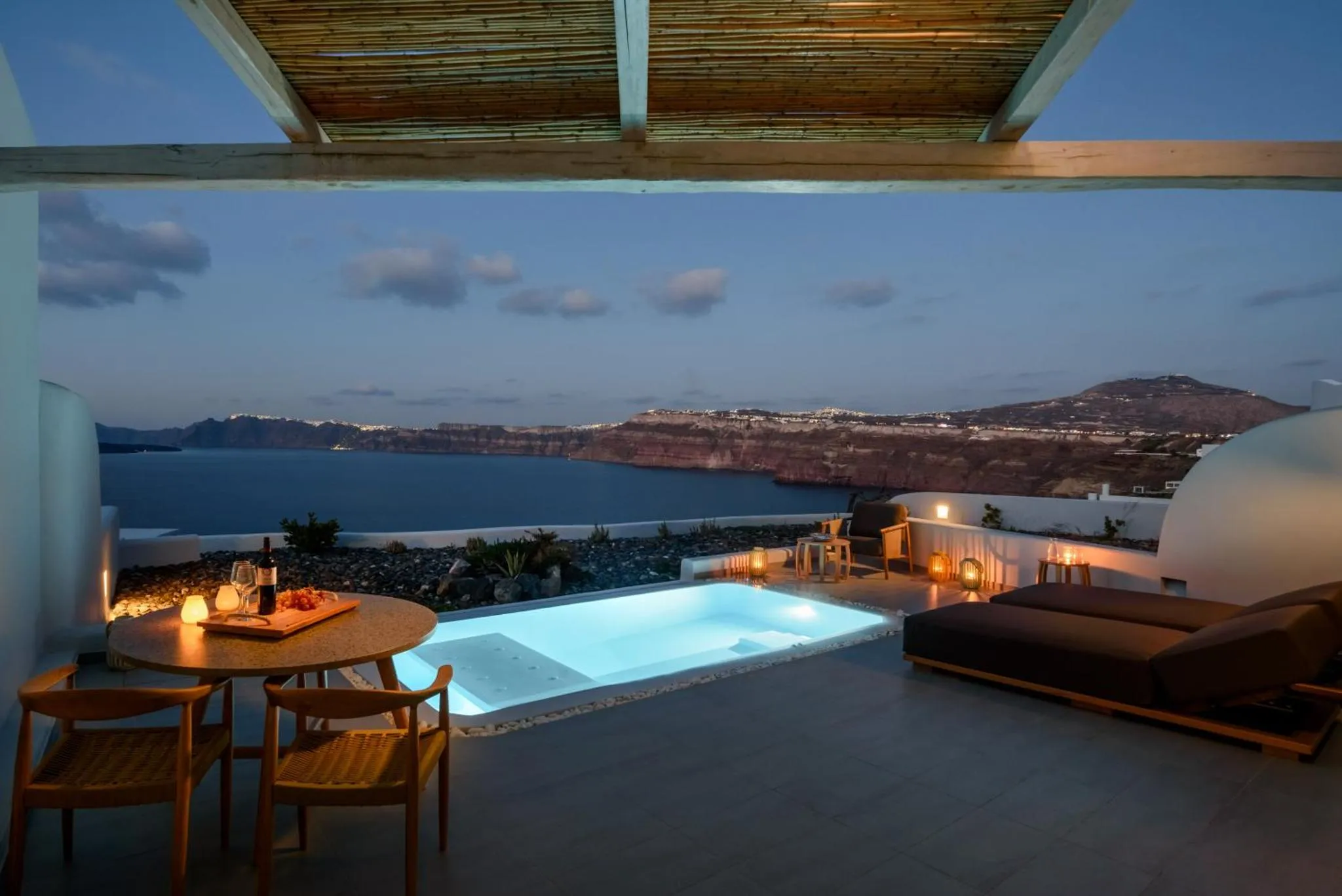 Une petite table ronde avec des boissons et une piscine hors sol offrent une vue sur la mer et les collines illuminées.