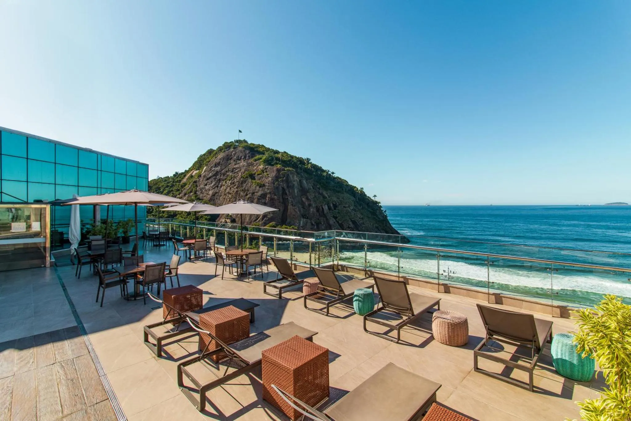 Terrasse aménagée avec transats et tables offrant une vue sur la mer et une colline.
