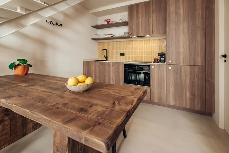 Une table en bois rustique est placée devant une cuisine moderne avec des armoires en bois et un carrelage jaune.