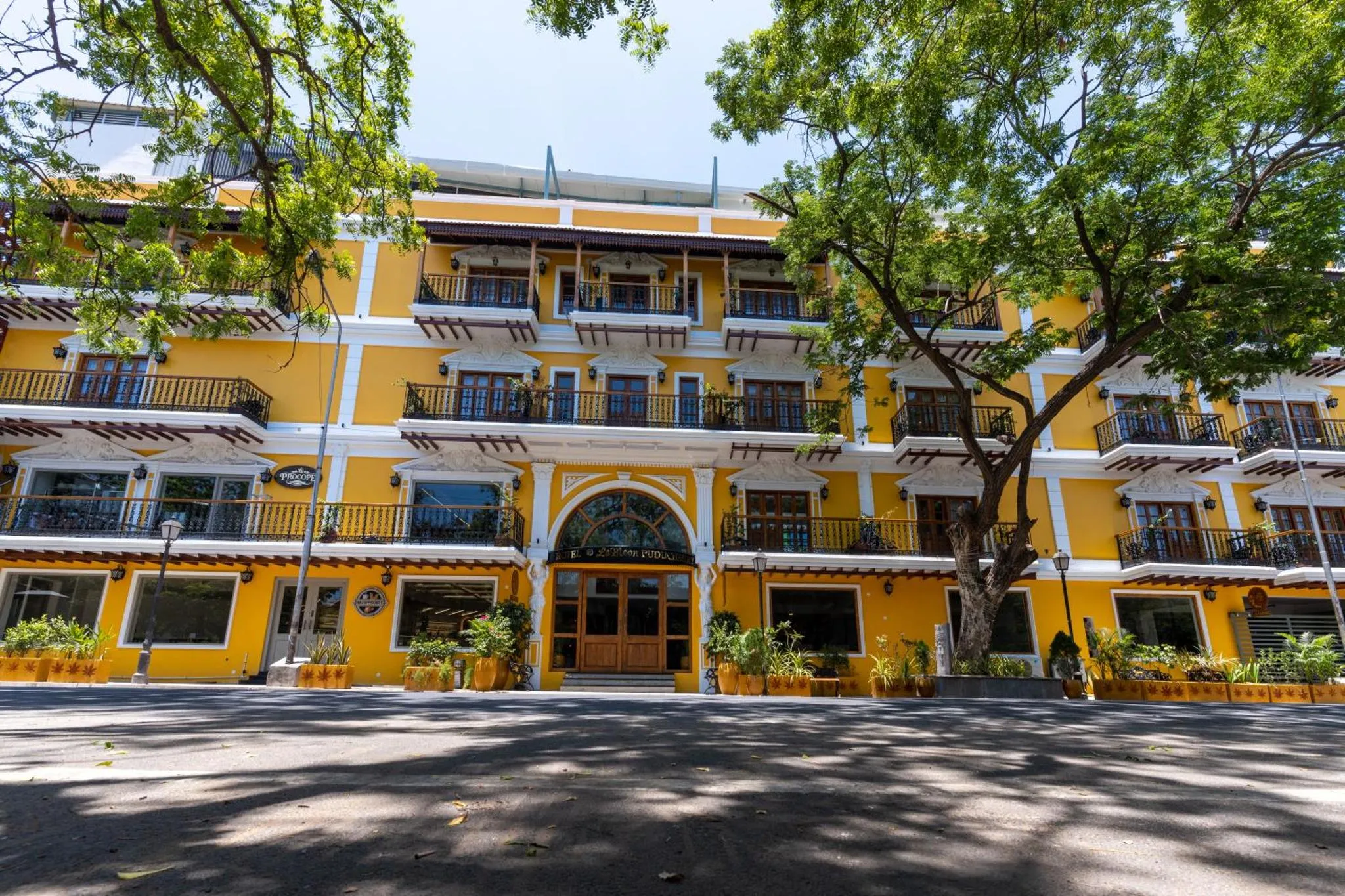 Facade jaune d'un bâtiment à balcons et arcades, ombragée par des arbres.