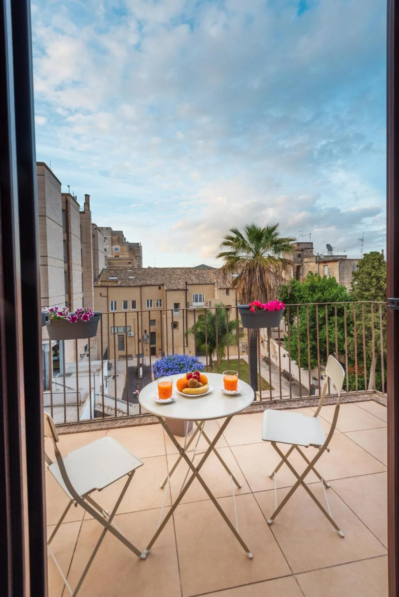 Une table ronde avec des verres et des fruits est posée sur un balcon fleuri.