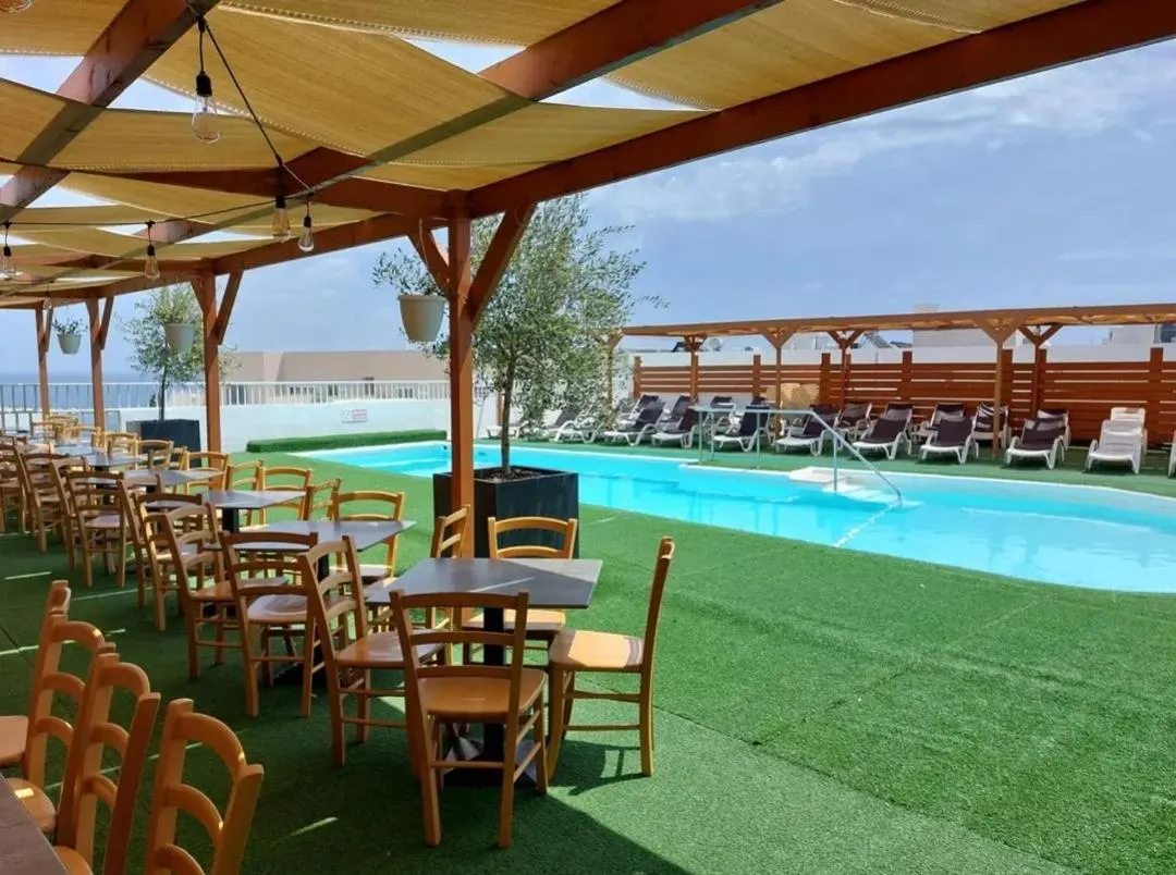 Terrasse ombragée avec tables et chaises donnant sur une piscine et un espace de détente.
