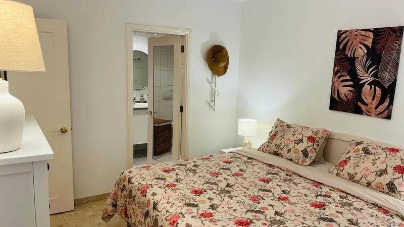 Une chambre lumineuse avec un lit recouvert d'une courtepointe fleurie et un accès à une salle de bain.