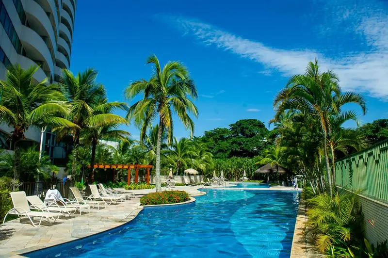 Une piscine rectangulaire bordée de chaises longues et de palmiers sous un ciel bleu.
