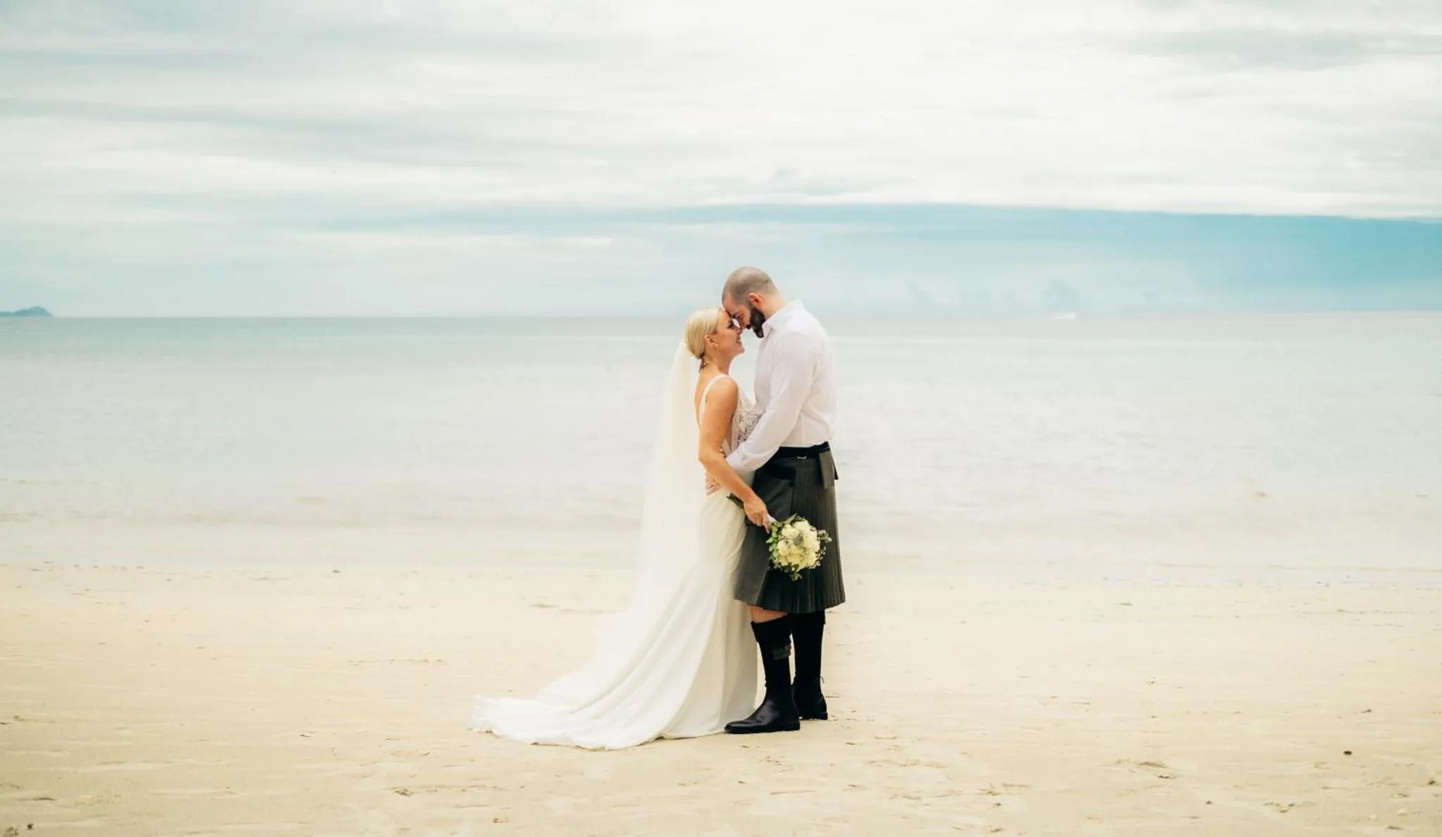 Un couple se tient sur une plage de sable clair, face à la mer.