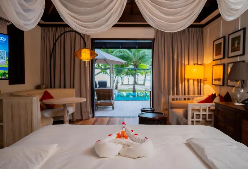 Une chambre lumineuse avec un lit décoré de serviettes et une vue sur une piscine et la plage.