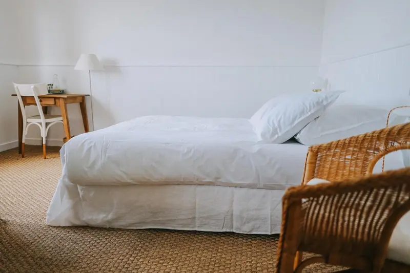 Une chambre lumineuse avec un lit recouvert de draps blancs et un bureau en bois.