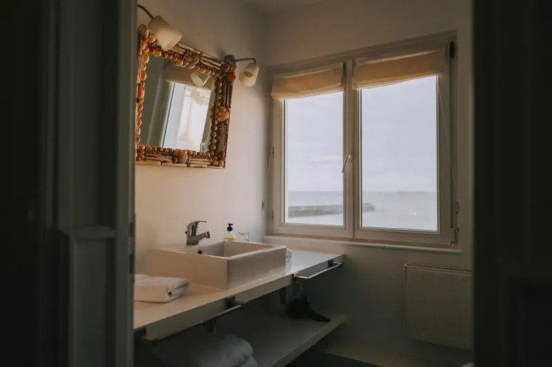 Une salle de bain lumineuse avec un miroir orné et vue sur la mer.