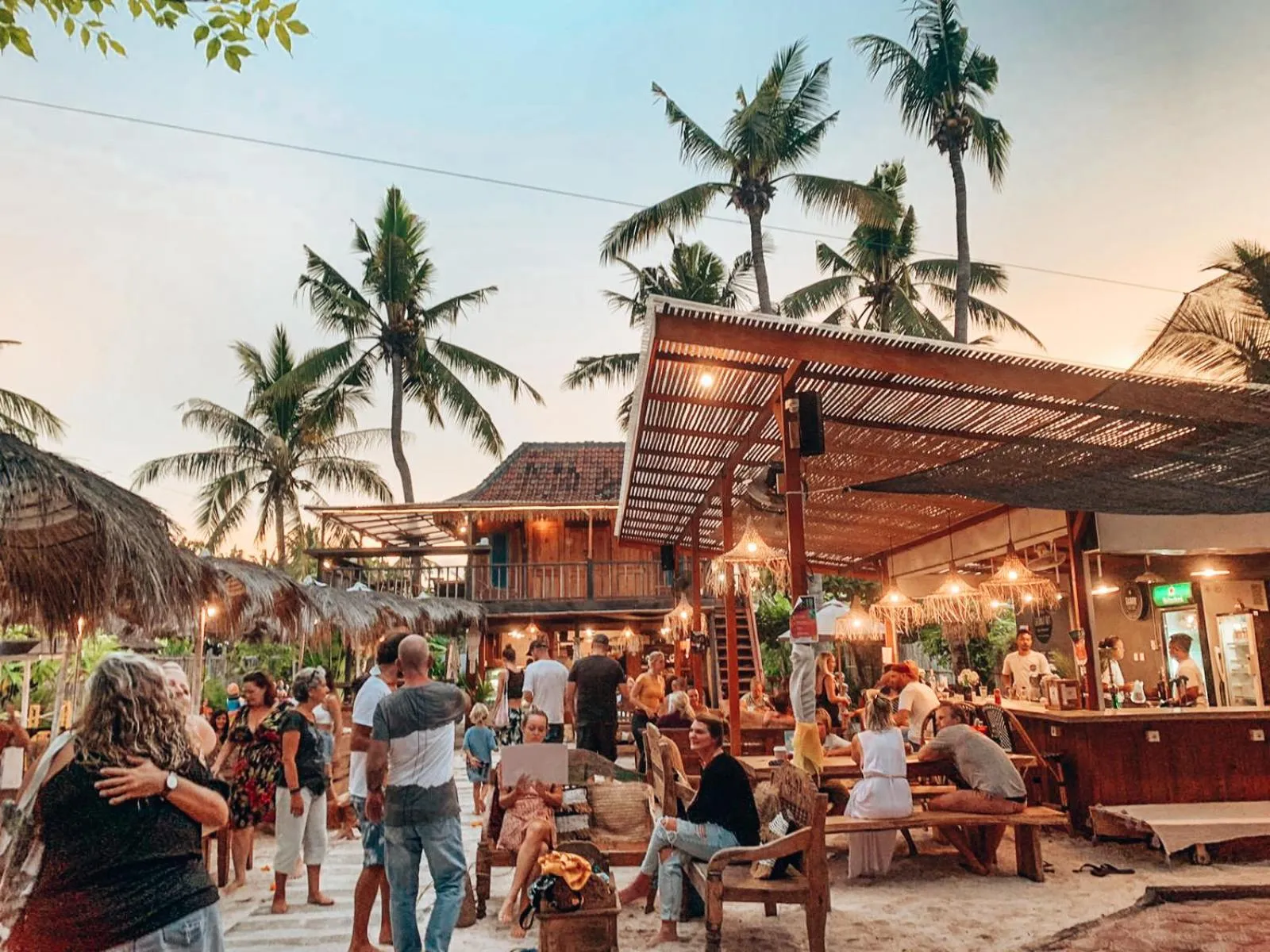 Une terrasse de restaurant animée avec des clients assis à des tables en bois sous un toit de chaume.