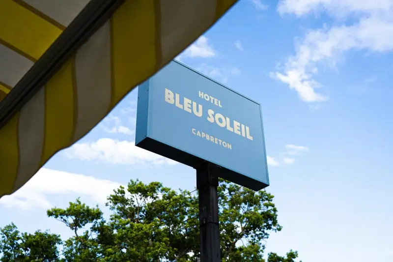 Panneau d'hôtel bleu et blanc sous un auvent jaune, avec des arbres verts en arrière-plan et un ciel bleu clair.