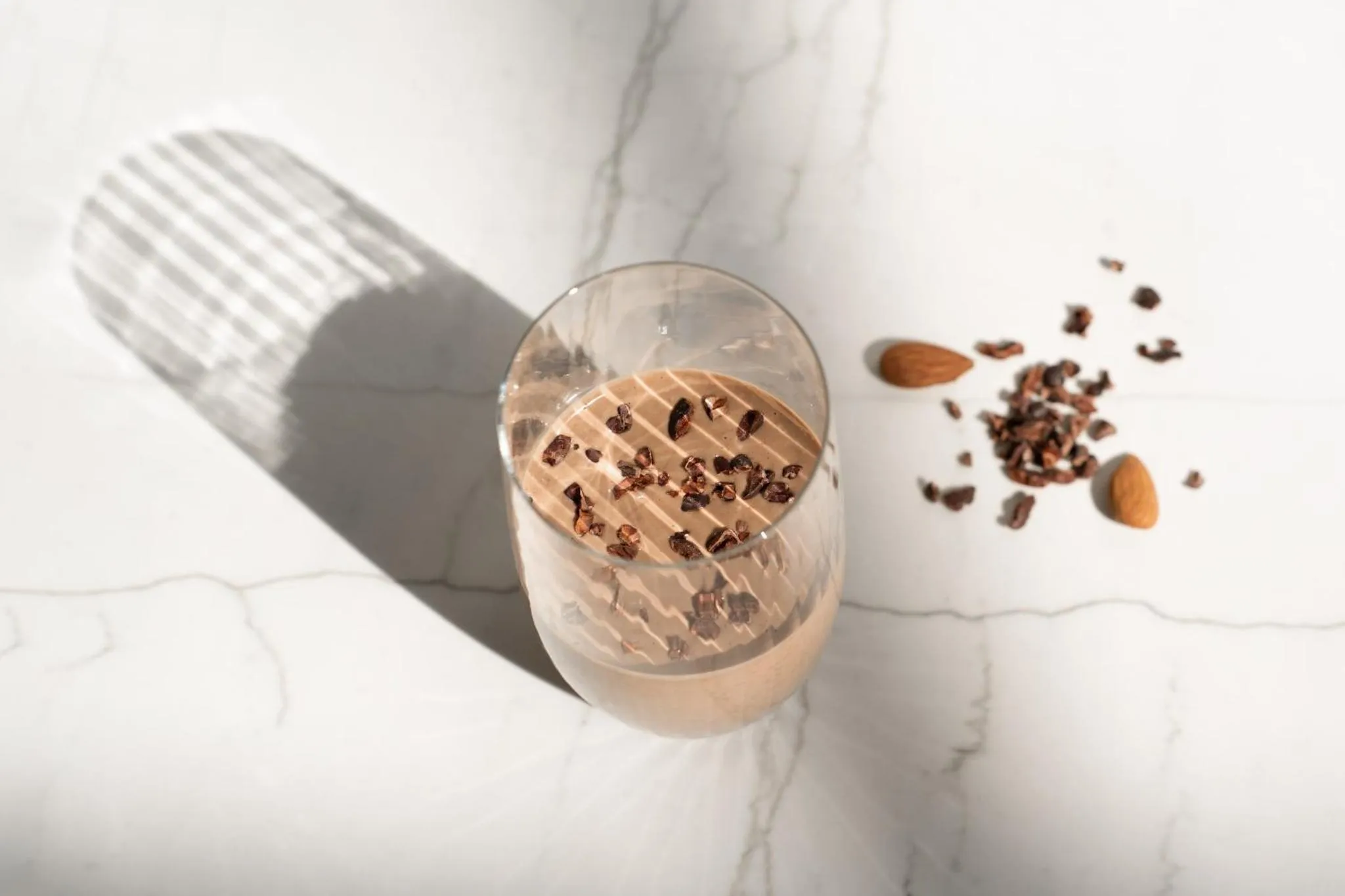 Un verre de boisson crémeuse est accompagné de quelques amandes et de morceaux de chocolat sur une surface marbrée.