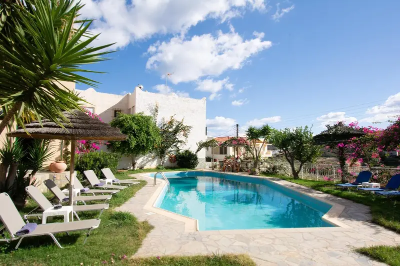 Transats alignés au bord d'une piscine ensoleillée, avec une végétation luxuriante et des bâtiments blancs en arrière-plan.